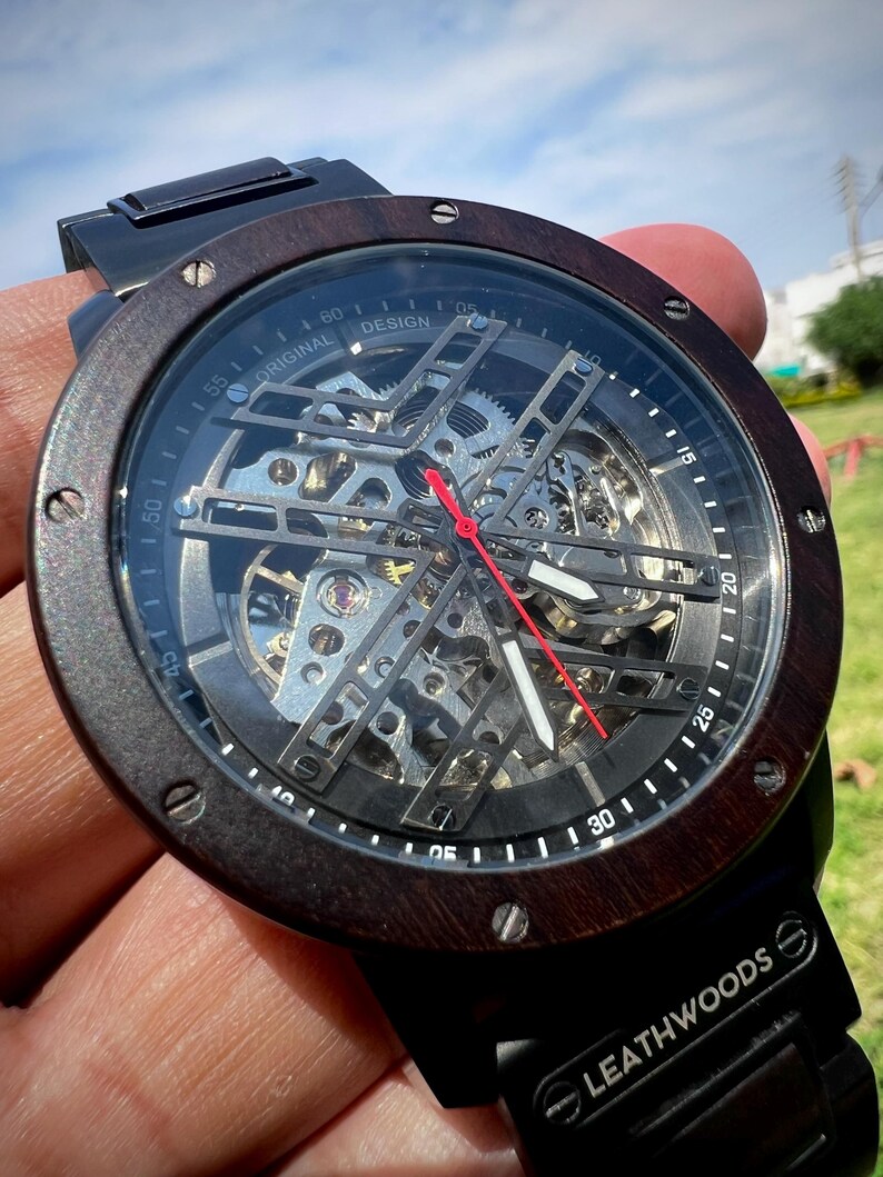 May include: A skeleton watch with a dark wood bezel and black band. The watch face reveals the intricate inner workings with silver gears and a red second hand. The watch is marked "ORIGINAL DESIGN" and "LEATHWOODS".
