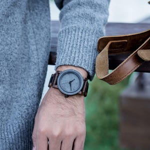 May include: A wristwatch with a dark gray face and brown wooden band is shown on a person's wrist. The watch has a minimalist design with simple hands. The band appears to be made of dark wood. The watch is a stylish accessory.