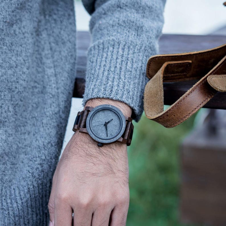 May include: A wristwatch with a dark gray face and brown wooden band is shown on a person's wrist. The watch has a minimalist design with simple hands. The band appears to be made of dark wood. The watch is a stylish accessory.