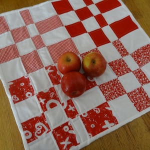 May include: A red and white checkered table runner with a red and white gingham pattern. Three red apples are on top of the runner.