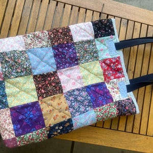 Può includere: Una borsa tote patchwork con una varietà di motivi floreali in quadrati rosa, blu, viola e rossi. La borsa ha una texture trapuntata e manici blu scuro. La borsa è su un tavolo di legno.