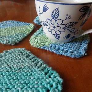 May include: Four crocheted coasters in shades of green and blue. The coasters are made with a textured stitch and are ideal for protecting surfaces from hot or cold drinks.