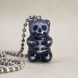 Op de afbeelding: Een donkerpaarse gummybeer hanger met een schedelontwerp, bevestigd aan een zilveren kogelketting. Het skelet van de beer is zichtbaar, wat een unieke en spookachtige esthetiek creëert. De ketting is een leuk accessoire.
