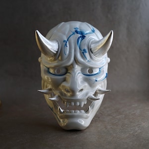 Könnte beinhalten: Eine weiße japanische Dämonenmaske aus Keramik mit silbernen Hörnern und Zähnen. Die Maske hat blaue dekorative Akzente und einen grimmigen Ausdruck. Ein dekorativer Artikel.