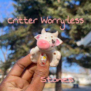 Op de afbeelding: Een kleine, witte keramische koeienfiguur met bruine vlekken, roze oren en een roze neus. De tekst "critter Worry-less Stones" staat in roze boven en onder het beeldje. De koe wordt vastgehouden in een hand met een bloemenontwerp op de nagel.