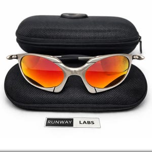 Op de afbeelding: Zilverkleurige zonnebril met rood-oranje glazen ligt op een zwarte etui. De zonnebril heeft een strak, modern design. De etui is open en toont de bril. De tekst "RUNWAY LABS" staat op een wit label.