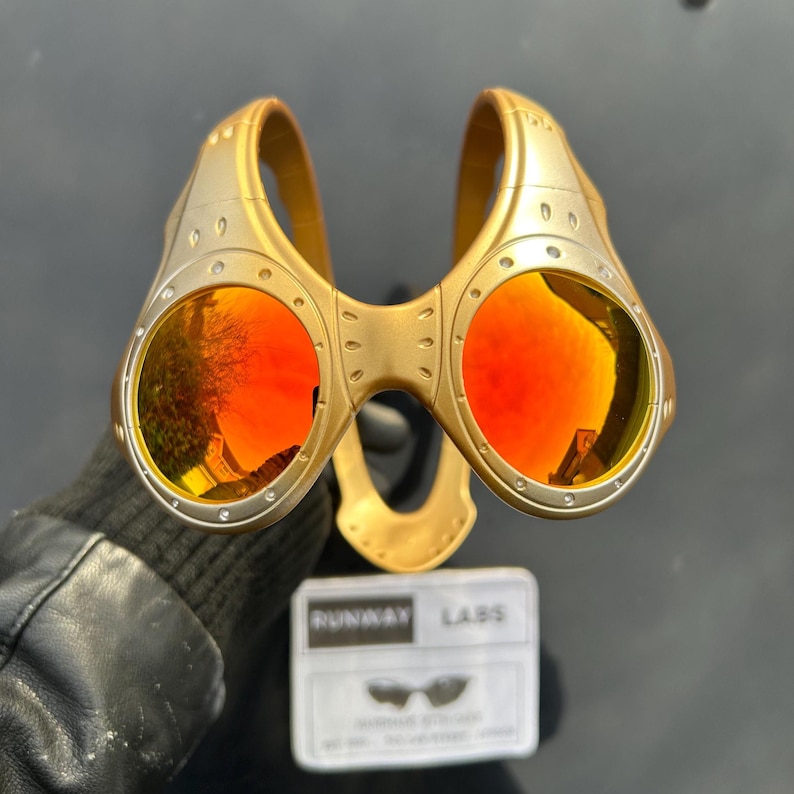 Op de afbeelding: Goudkleurige steampunk-zonnebril met ronde, oranje getinte glazen. Het montuur heeft een geklonken ontwerp en een brug die de twee glazen verbindt. Een klein wit label met de tekst "RUNWAY LABS" is zichtbaar.