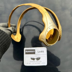 Op de afbeelding: Goudkleurige zonnebril met een uniek, futuristisch design. De bril heeft een gebogen montuur en ronde glazen met een reflecterende, gouden tint. Een wit label met de tekst "RUNWAY LABS" is ook aanwezig.