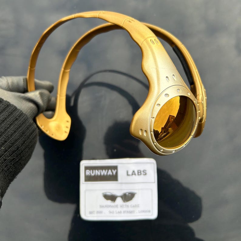 Op de afbeelding: Goudkleurige zonnebril met een uniek, futuristisch design. De bril heeft een gebogen montuur en ronde glazen met een reflecterende, gouden tint. Een wit label met de tekst "RUNWAY LABS" is ook aanwezig.