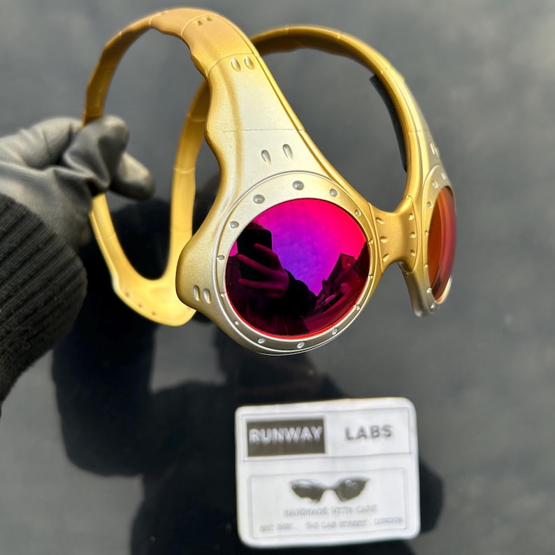 Op de afbeelding: Goudkleurige en zilverkleurige zonnebril met felroze glazen. De zonnebril heeft een uniek, industrieel ontwerp met zichtbare schroeven en een gebogen montuur. Een kleine witte kaart met de tekst "RUNWAY LABS" is ook aanwezig.