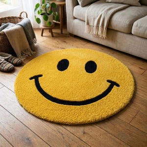 Pu&ograve; includere: Un tappeto rotondo giallo con un grande disegno di faccina sorridente nera. Il tappeto &egrave; realizzato in un materiale morbido e strutturato ed &egrave; posizionato su un pavimento in legno. Il diametro del tappeto &egrave; di circa 122 cm.