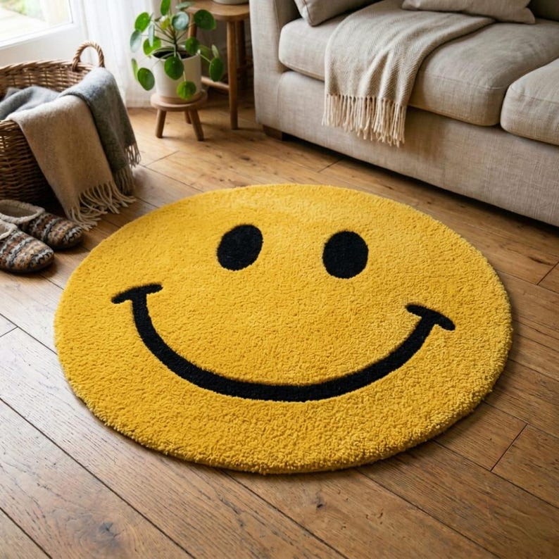 Pu&ograve; includere: Un tappeto rotondo giallo con un grande disegno di faccina sorridente nera. Il tappeto &egrave; realizzato in un materiale morbido e strutturato ed &egrave; posizionato su un pavimento in legno. Il diametro del tappeto &egrave; di circa 122 cm.