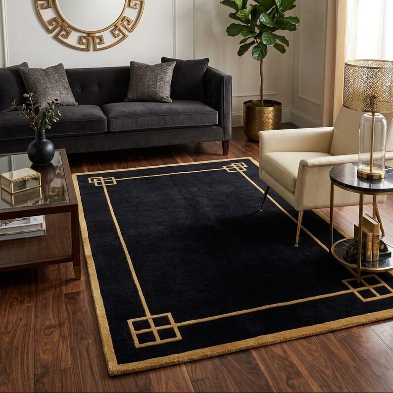 May include: A black rectangular rug with a gold geometric border and corner patterns. The rug is on a dark wood floor in a living room, with a sofa, armchair, and side table. The rug's dimensions are in centimetres.