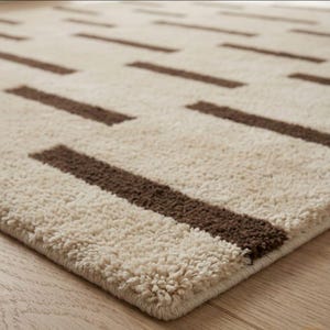 Puede incluir: Una alfombra rectangular beige con un patr&oacute;n repetitivo de gruesas rayas rectangulares de color marr&oacute;n oscuro. La alfombra tiene una superficie suave y texturizada y est&aacute; colocada sobre un suelo de madera. El dise&ntilde;o de la alfombra a&ntilde;ade un toque moderno a cualquier habitaci&oacute;n.