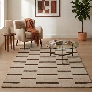 Puede incluir: Alfombra rectangular beige con un patr&oacute;n repetido de rayas horizontales marr&oacute;n oscuro. La alfombra est&aacute; en un sal&oacute;n con una mesa de centro redonda de cristal, un sill&oacute;n beige y una obra de arte enmarcada en la pared.
