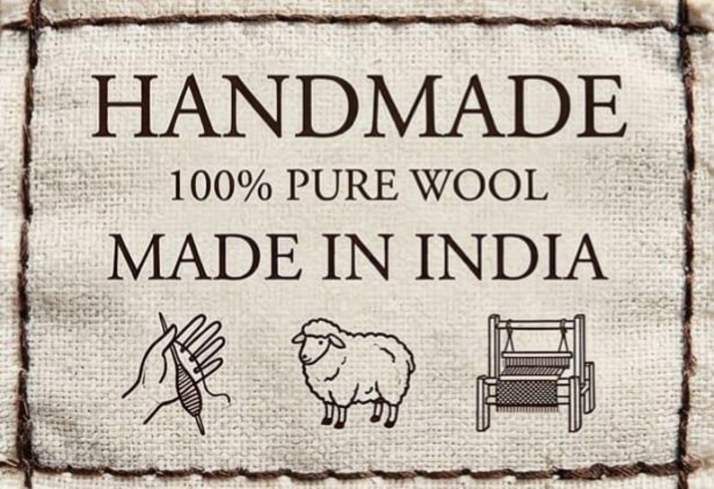 May include: A cream-colored fabric label with the words "HANDMADE", "100% PURE WOOL", and "MADE IN INDIA" in dark brown text. The label features illustrations of a hand with yarn, a sheep, and a loom.