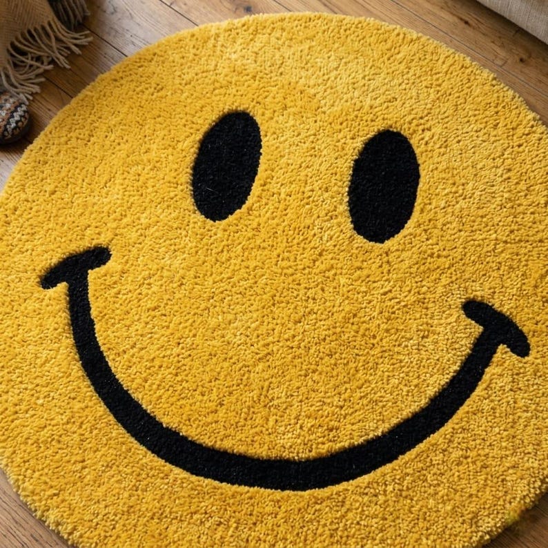 Pu&ograve; includere: Un tappeto rotondo giallo con un design a faccina sorridente. Il tappeto presenta una superficie strutturata e una faccina sorridente nera con due occhi ovali e una bocca curva. Il tappeto &egrave; progettato per l'arredamento della casa.