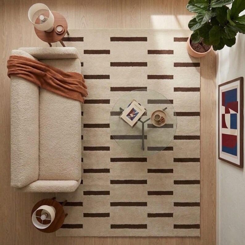 Puede incluir: Vista a&eacute;rea de una sala de estar. Un sof&aacute; color crema con una manta color &oacute;xido se encuentra junto a una alfombra con rayas marrones. Una mesa de centro de cristal tiene un libro y una bandeja peque&ntilde;a. Tambi&eacute;n se ve una obra de arte enmarcada y una planta en maceta.