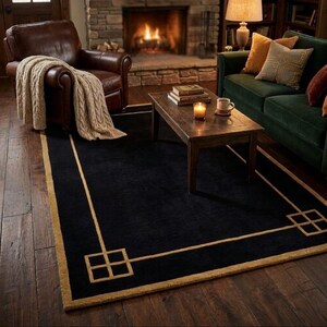 May include: A black rectangular rug with a gold border and geometric corner detailing. The rug is positioned on a wooden floor, with a fireplace and furniture in the background, creating a cozy living room atmosphere.