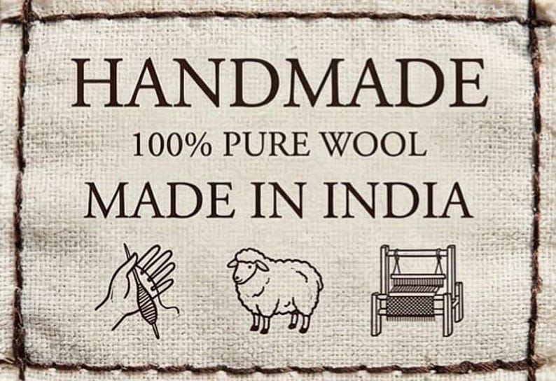 May include: A cream fabric label with the text "HANDMADE", "100% PURE WOOL", and "MADE IN INDIA" in dark brown. The label includes illustrations of a hand with yarn, a sheep, and a loom.