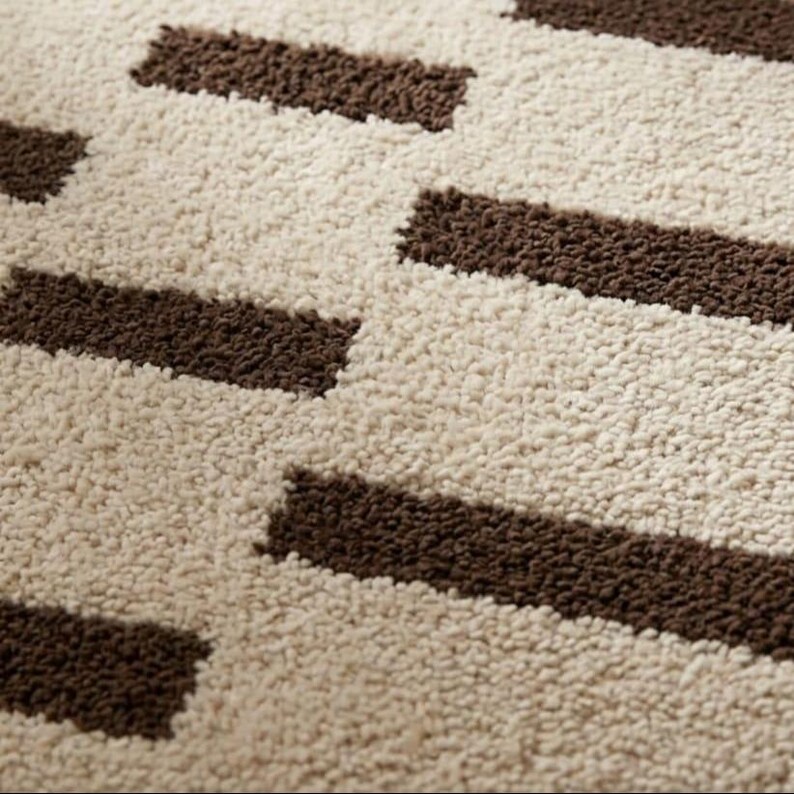 Puede incluir: Primer plano de una alfombra beige y marr&oacute;n estampada. La alfombra tiene una superficie texturizada con formas rectangulares marrones alternas sobre un fondo beige. El dise&ntilde;o crea un patr&oacute;n geom&eacute;trico.
