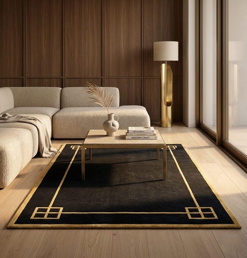 May include: A black rectangular rug with a gold geometric border and accents. The rug is placed in a living room setting with a beige sectional sofa, a wooden coffee table, and a gold floor lamp.