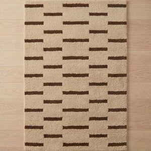 Puede incluir: Alfombra rectangular beige con un patr&oacute;n repetitivo de rayas horizontales marr&oacute;n oscuro. La alfombra tiene un aspecto suave y texturizado, adecuada para un ba&ntilde;o o dormitorio. El dise&ntilde;o es simple y moderno, ofreciendo una paleta de colores neutros.