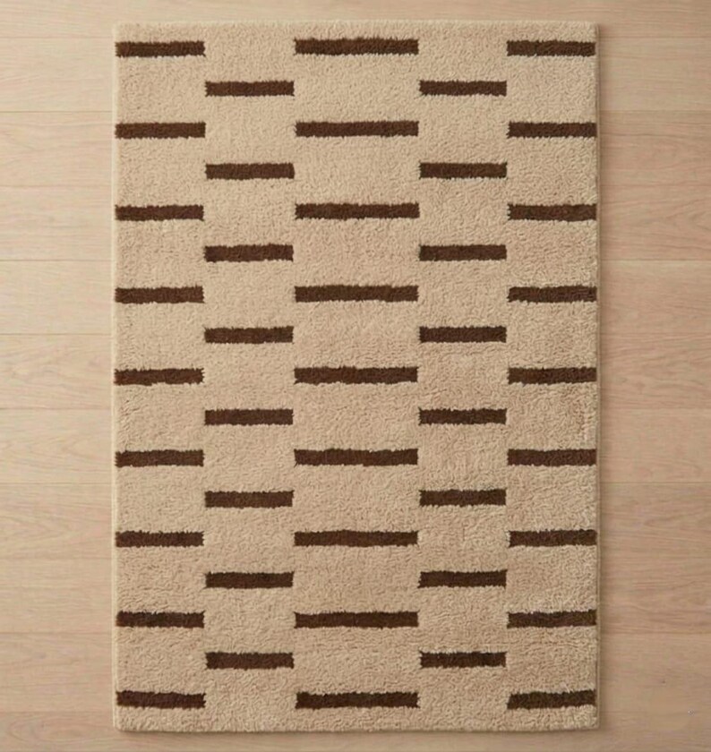 Puede incluir: Alfombra rectangular beige con un patr&oacute;n repetitivo de rayas horizontales marr&oacute;n oscuro. La alfombra tiene un aspecto suave y texturizado, adecuada para un ba&ntilde;o o dormitorio. El dise&ntilde;o es simple y moderno, ofreciendo una paleta de colores neutros.