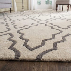 May include: A cream-colored area rug with a geometric pattern in gray. The rug has a soft, textured appearance and is placed on a dark wood floor. The pattern is a repeating design of interlocking shapes.