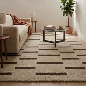 Puede incluir: Una alfombra rectangular beige con un patr&oacute;n repetido de rayas horizontales marr&oacute;n oscuro. La alfombra est&aacute; colocada en un sal&oacute;n con un sof&aacute;, una mesa de centro y una planta en maceta. El dise&ntilde;o de la alfombra a&ntilde;ade un toque moderno a la habitaci&oacute;n.