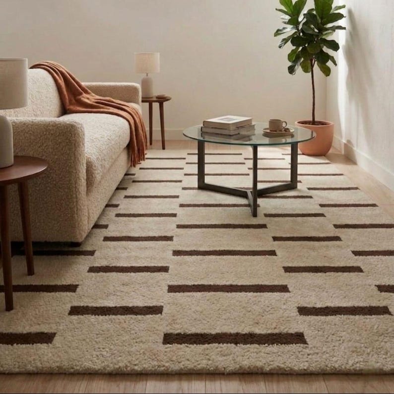 Puede incluir: Una alfombra rectangular beige con un patr&oacute;n repetido de rayas horizontales marr&oacute;n oscuro. La alfombra est&aacute; colocada en un sal&oacute;n con un sof&aacute;, una mesa de centro y una planta en maceta. El dise&ntilde;o de la alfombra a&ntilde;ade un toque moderno a la habitaci&oacute;n.