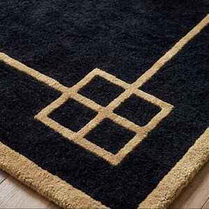 May include: A close-up of a black rug with a geometric design. The rug features a square pattern in a tan colour, with a tan border. The rug is made of a soft, textured material.