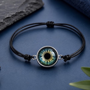 Op de afbeelding: Een zwarte koordarmband met een centrale ronde bedel met een realistisch blauw en geel menselijk irisontwerp onder een glazen koepel, gevat in een zilverkleurige metalen zetting met verstelbare schuifknopen voor een goede pasvorm.
