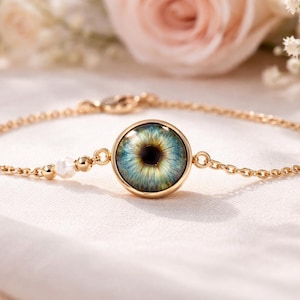 Peut inclure: Un bracelet doré orné d'un pendentif en forme d'œil détaillé. Le médaillon rond présente un iris réaliste dans des tons de bleu et de vert. La chaîne fine comprend de petites perles blanches et un fermoir, créant un bijou élégant.