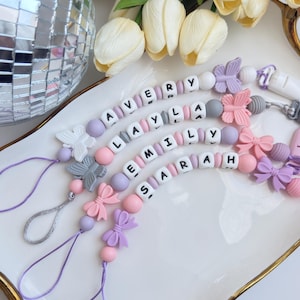 May include: Four personalised pacifier clips with names spelled out in white letter beads. The clips feature pastel pink, lilac, and grey beads, butterfly and bow accents, and a loop for attachment. White tulips and a disco ball are in the background.