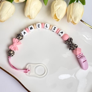 Op de afbeelding: Een speenkoord met een roze koord, een doorzichtige siliconen ring en een reeks kralen waaronder witte letterkralen die AMELIA spellen, roze ronde kralen, roze strikkralen en kralen met luipaardprint, afgewerkt met een effen roze clip.