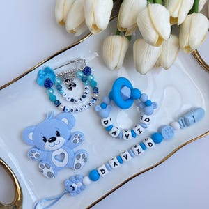 Puede incluir: Un conjunto de accesorios para bebés en tonos azules, que incluyen un clip para chupete, un chupete, un imperdible y un mordedor con forma de oso de peluche. El clip del chupete deletrea el nombre "DAVID" con cuentas de letras blancas. En una bandeja blanca.