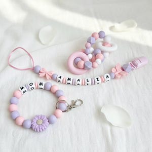 以下が含まれることがあります： パステルピンクとラベンダー色のベビーアクセサリーセット。セットには、「AMALIA」という名前の付いたおしゃぶりクリップ、歯固め、そして「MOM」という文字が入ったブレスレットが含まれています。ブレスレットには花のチャームと留め金が付いています。おしゃぶりクリップには蝶のチャームが付いています。