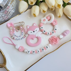 May include: A collection of baby accessories in pink and white, including a pacifier clip, a teething ring, and a fox-shaped teether. The pacifier clip and teething ring have the name "CHLOE" spelled out in letter beads. A decorative pin is also included.