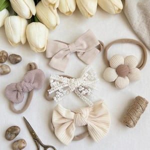May include: A collection of baby headbands in beige and cream hues, featuring bow and flower designs. The headbands are displayed with white tulips, small stones, scissors, and twine, creating a delicate, feminine arrangement.