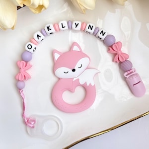 May include: A pink fox-shaped teether and pacifier clip with the name "Oaklynn" spelled out in white letter beads. The clip features pink bows and beads. The teether is made of soft silicone.