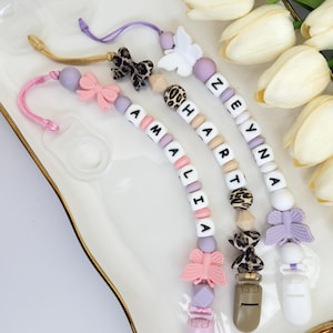May include: Three personalised baby dummy clips with names spelled out in white letter beads. Each clip features a different design, including bows, butterflies, and leopard print accents. The clips are in shades of pink, purple, and beige.