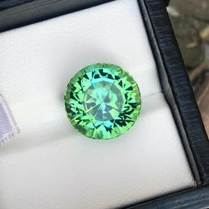 May include: A round, faceted gemstone with a vibrant green hue, displayed in a black box with a white interior. The gem's facets reflect light, creating a sparkling effect. The gem is a striking piece of jewelry.