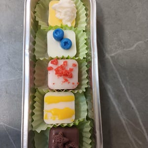 Puede incluir: Vista cenital de un recipiente metálico rectangular con cinco pequeños bombones cuadrados en moldes de papel verde. Los bombones están decorados con varios ingredientes, como nata montada, arándanos y virutas. Los bombones están dispuestos en fila.