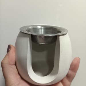 Puede incluir: Un portavelas blanco en forma de huevo con un recorte en forma de U. Cuenta con una copa de metal plateado en la parte superior para sostener una vela. El portavelas se sostiene en una mano.