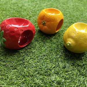 Puede incluir: Tres quemadores de aceite de cerámica con forma de fruta. Una fresa roja con hojas verdes, una naranja y un limón amarillo. Cada uno tiene un agujero para una vela de té y un cuenco para aceites esenciales. Los quemadores están sobre una superficie verde y herbosa.