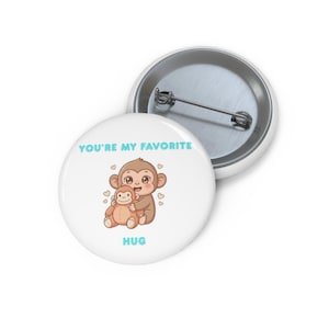 Könnte beinhalten: Ein weißer Ansteckbutton mit einer Cartoon-Illustration von zwei sich umarmenden Affen. Der Text "YOU'RE MY FAVORITE" und "HUG" sind hellblau. Der Button hat eine Metallrückseite.