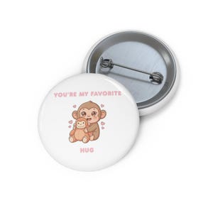 Könnte beinhalten: Ein weißer Button mit einer Cartoon-Illustration von zwei sich umarmenden Affen. Der Text "YOU'RE MY FAVORITE" und "HUG" ist in Rosa gehalten. Der Button hat eine Metallrückseite und eine Anstecknadel.