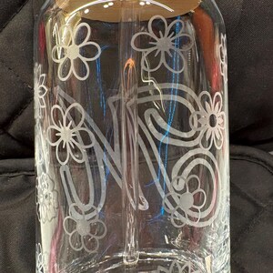 May include: A clear glass tumbler with a bamboo lid and a glass straw. The exterior features a white etched design of multiple flowers and a large letter N in the center. The glass has a cylindrical shape with a rounded base and a wide opening.