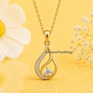 Pode incluir: Um colar de tom dourado com um pingente em forma de gota e design curvo. Um lado do pingente é forrado com pequenas pedras claras e uma pedra clara maior está fixada no centro. O texto GemstoneTownDesign é visível na imagem.